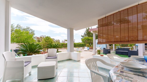 A covered outdoor seating area with white wicker furniture, a tiled floor, and a view of greenery and buildings in the background. - Sardinian Apartment with Private Terrace 3 Minutes’ Walk from the Beach (Orosei)