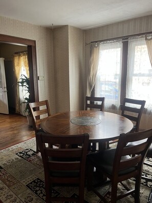 Une table de salle à manger ronde en bois, accompagnée de quatre chaises, un tapis à motifs et une vue sur un bâtiment visible à travers les rideaux.