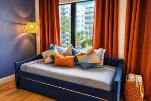 Een bed met meerdere kussens, een lamp en uitzicht op gebouwen door een raam.