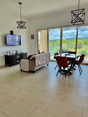 A spacious living area with a sofa, dining table, and chairs, a TV, and a view of a lush landscape through large windows. - Dream Getaway | Pool View + Resort Beach Access (María Chiquita)