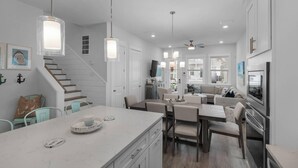 A modern kitchen with a central island, dining area with chairs, a staircase, and a living room with a sofa. - Emerald Escape 30A (Santa Rosa Beach)