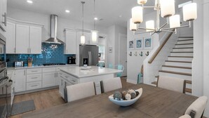 A modern kitchen with white cabinets, a blue backsplash, stainless steel appliances, and a dining area with a wooden table and chairs. - Emerald Escape 30A (Santa Rosa Beach)