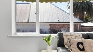 Ein Zimmer mit einem großen Fenster, einem schwarzen Ledersofa, einem kleinen Holztisch mit einer Pflanze und einer weißen Wand.