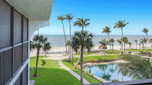 Un balcón con vista a una playa, palmeras, una piscina y una estructura de techo de paja.