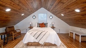 Ein gemütliches Schlafzimmer im Dachgeschoss mit schrägen Wänden, Holzbalken, einem Bett mit weißer Steppdecke, zwei Nachttischen mit Lampen, einem Sessel und einem Fenster mit Jalousien.