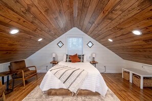 A cozy attic bedroom with a sloped ceiling, wooden beams, a bed with a white quilt, two bedside tables with lamps, a chair, and a window with blinds. - Dogwood Den Treehouse Cabin in Shirley, AR at Greers Ferry Lake (Edgemont)