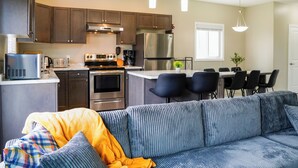 A modern kitchen with stainless steel appliances, a dining area with black chairs, and a blue sofa with a yellow blanket.