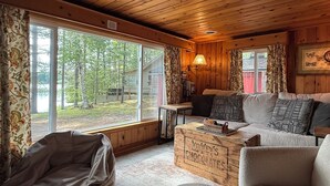 Ein Ferienhausinnenraum mit Holzbalkendecke, einer Couch, einem Sitzsack, einem Tisch mit Schachbrett und einem großen Fenster mit Blick auf ein bewaldetes Gebiet.
