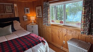 Ein Schlafzimmer in einer Holzhütte mit einem Bett, einem Nachttisch, einer Lampe, einem Fenster mit Vorhängen und Blick auf einen See.