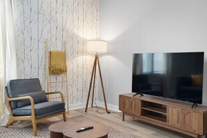 Ein Wohnzimmer mit Fernseher, Holzschrank, Sessel, Lampe und Wandpapier mit Muster.