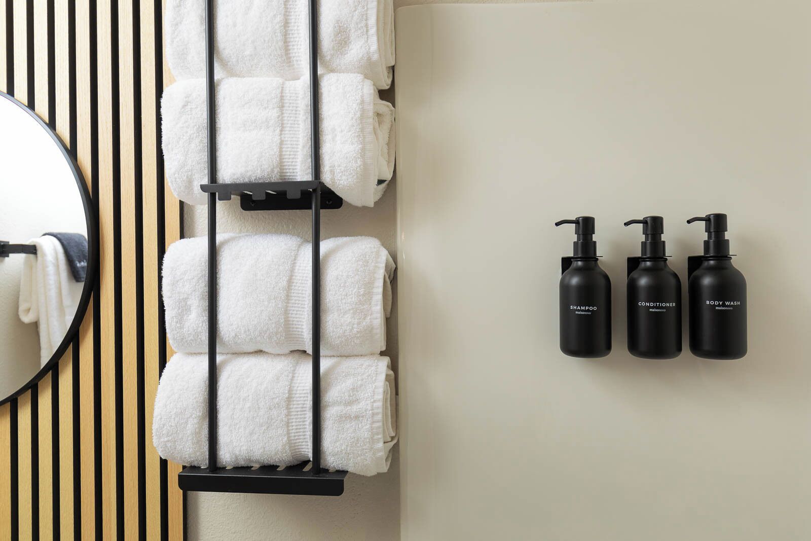 A bathroom with a towel rack holding several towels, a mirror, and three black bottles labeled shampoo, conditioner, and body wash.