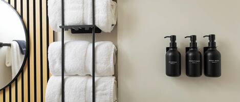 A bathroom with a towel rack holding several towels, a mirror, and three black bottles labeled shampoo, conditioner, and body wash.