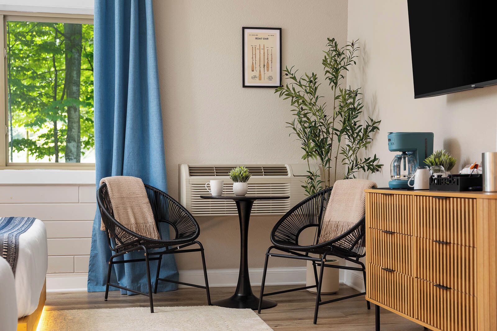 Ein modernes Zimmer mit einem Bett, zwei schwarzen Korbstühlen, einem kleinen Tisch mit Pflanze, einem an der Wand montierten Fernseher und einer Heizung.