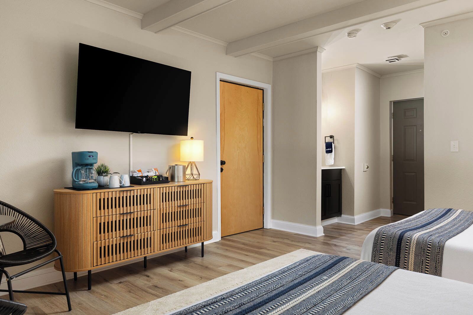 Ein Hotelzimmer mit Flachbildfernseher, einer Holzkonsole mit Kaffeemaschine, einer Lampe und einem Bett mit gestreifter Decke.