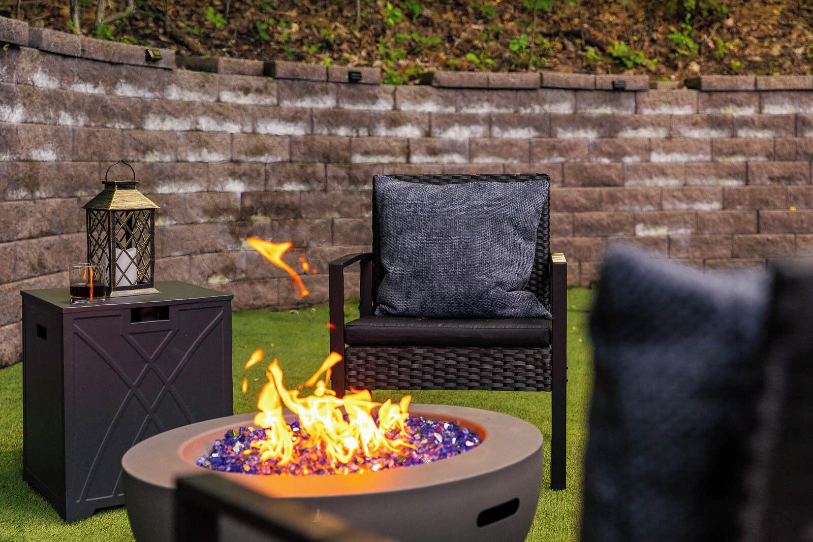 A fire pit with flames, a chair with a cushion, and a lantern on a stand.