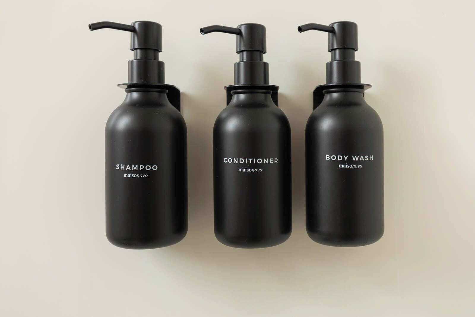 Trois flacons noirs dotés de pompes doseuses, portant les mentions « SHAMPOO », « APRÈS-SHAMPOO » et « GEL DOUCHE », et provenant de la marque « maisonovo ».