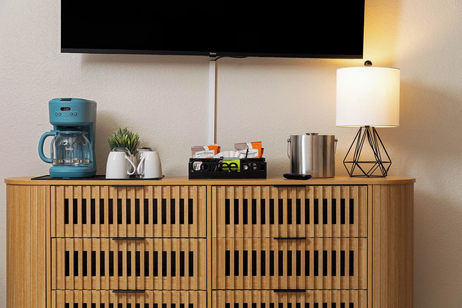 A wooden cabinet with a television mounted above it, a coffee maker, a kettle, a lamp, and a small plant.