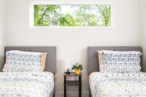 Quarto com duas camas, uma mesinha com uma planta e uma janela com vista para árvores.