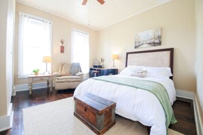 Une chambre à coucher avec un lit, une chaise, une table de chevet, une lampe, un tableau et une fenêtre.