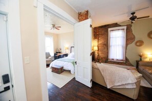 A bedroom with a bed, a desk, a chair, a window, and a brick wall. - Charming 1800`s Tiny Cottage | Walk to Lucas Oil! (Indianapolis)