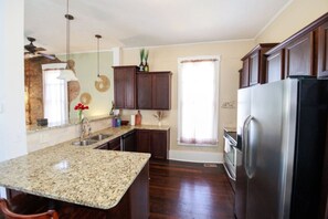 A kitchen with granite countertops, dark wooden cabinets, a stainless steel refrigerator, and a window with curtains. - Charming 1800`s Tiny Cottage | Walk to Lucas Oil! (Indianapolis)