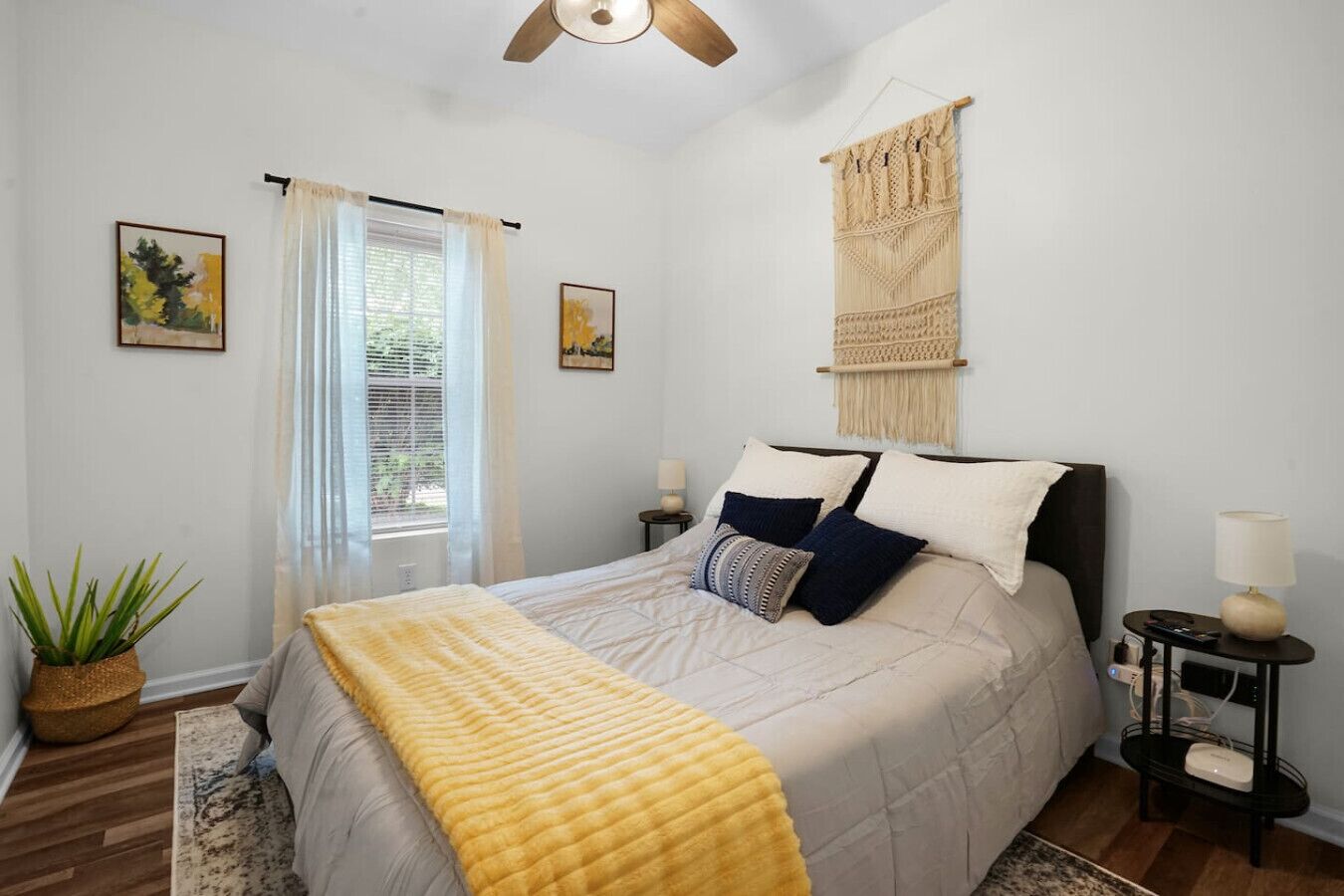 Un dormitorio con cama, mesitas de noche, ventilador de techo, ventana con cortinas y decoración en la pared.