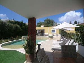 A poolside area with a brick pillar, lounge chairs, and a pergola with an umbrella.