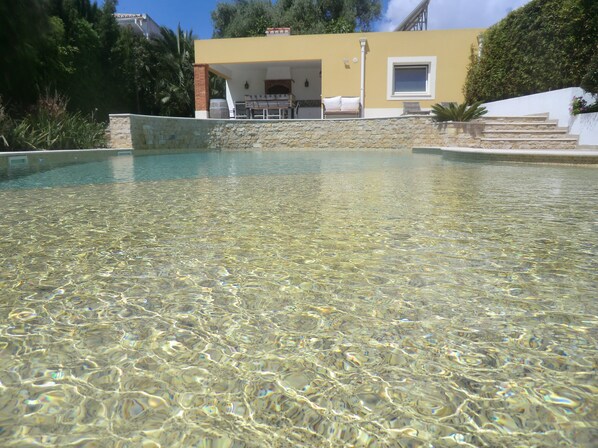 A clear pool with a stone wall, a yellow building with a patio, and steps leading up to it.