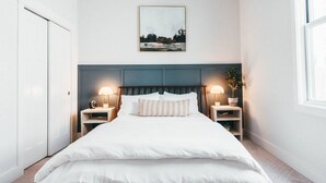 Een net opgemaakt bed met witte lakens, twee nachtkastjes met lampen, een hoofdbord en een ingelijst foto aan de muur.