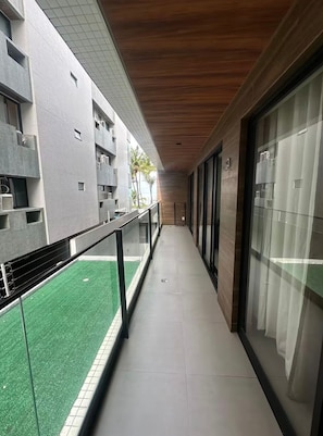 A corridor with a glass railing, a green artificial lawn, and a building with a tiled exterior and wooden ceiling. - Altamare 109 - Cozy beachfront apartment on Caribessa Beach (João Pessoa)