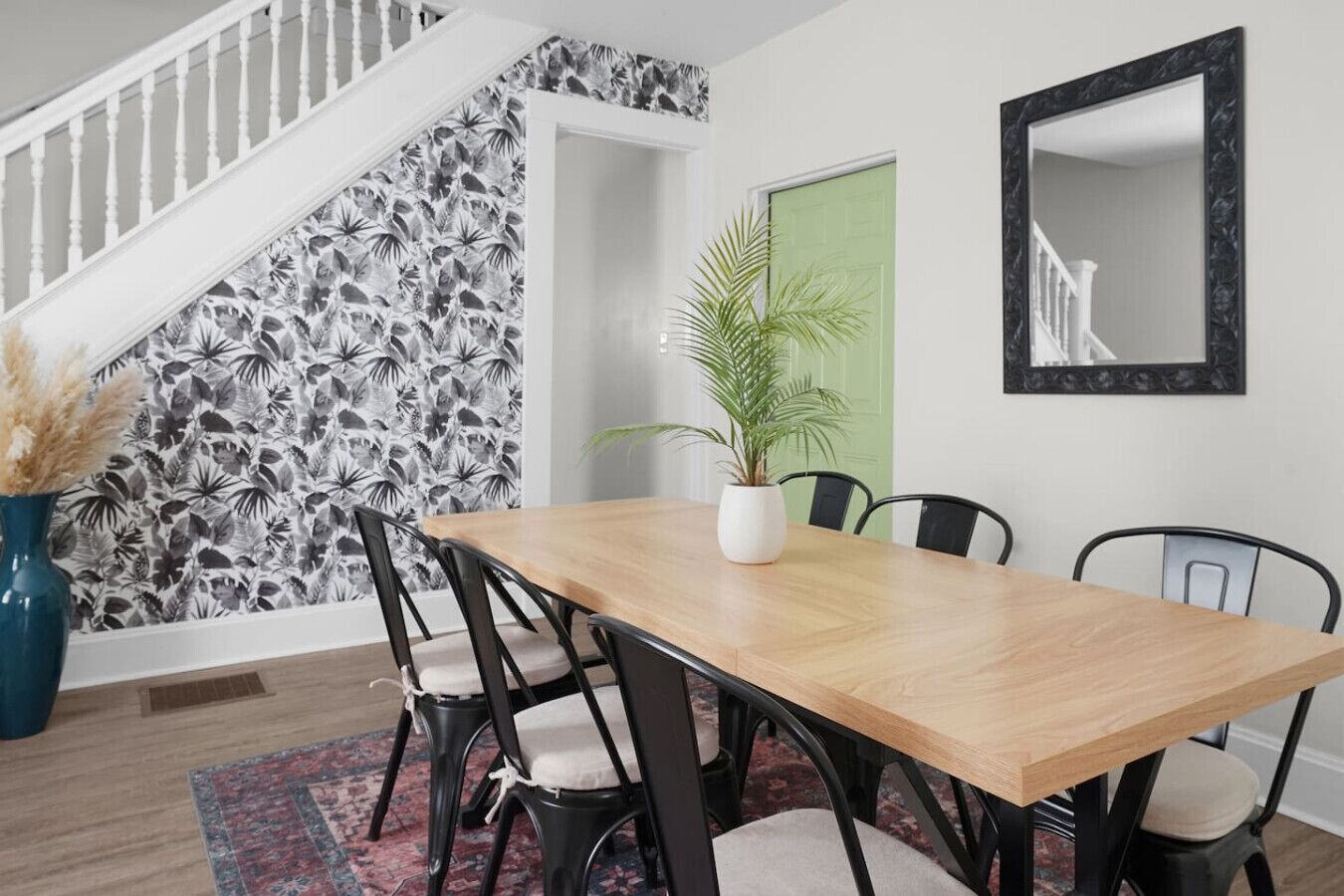 Un coin repas comprenant une table en bois, quatre chaises, une plante en pot, un miroir et un escalier dont les murs sont recouverts d’un papier peint à motifs floraux.