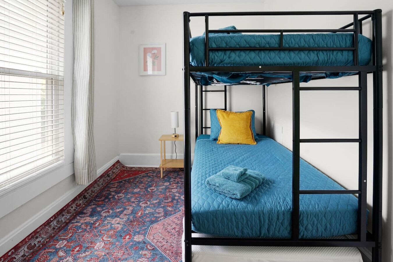 A bunk bed with a blue mattress and a yellow pillow, a small wooden nightstand, a red and blue patterned rug, and a framed picture on the wall.