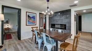 Un comedor con una mesa de madera, sillas azul claro, una araña, una chimenea y un cuadro enmarcado en la pared.