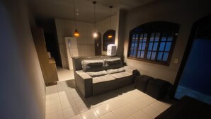 A living room with a sofa, a kitchen area, and a window with bars. - Beach house / Suarão - Itanhaém, 7 minutes walk from the beach. (Itanhaém)