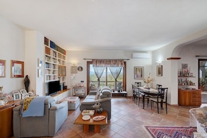 A living room with a sofa, a coffee table, a dining table with chairs, a bookshelf, and a television.