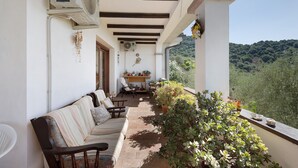 Un espacio de estar al aire libre cubierto con un sofá y sillones, una mesa con plantas en macetas y vistas a una ladera boscosa.
