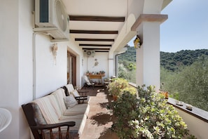 A covered outdoor seating area with a sofa and chairs, a table with potted plants, and a view of a forested hillside.