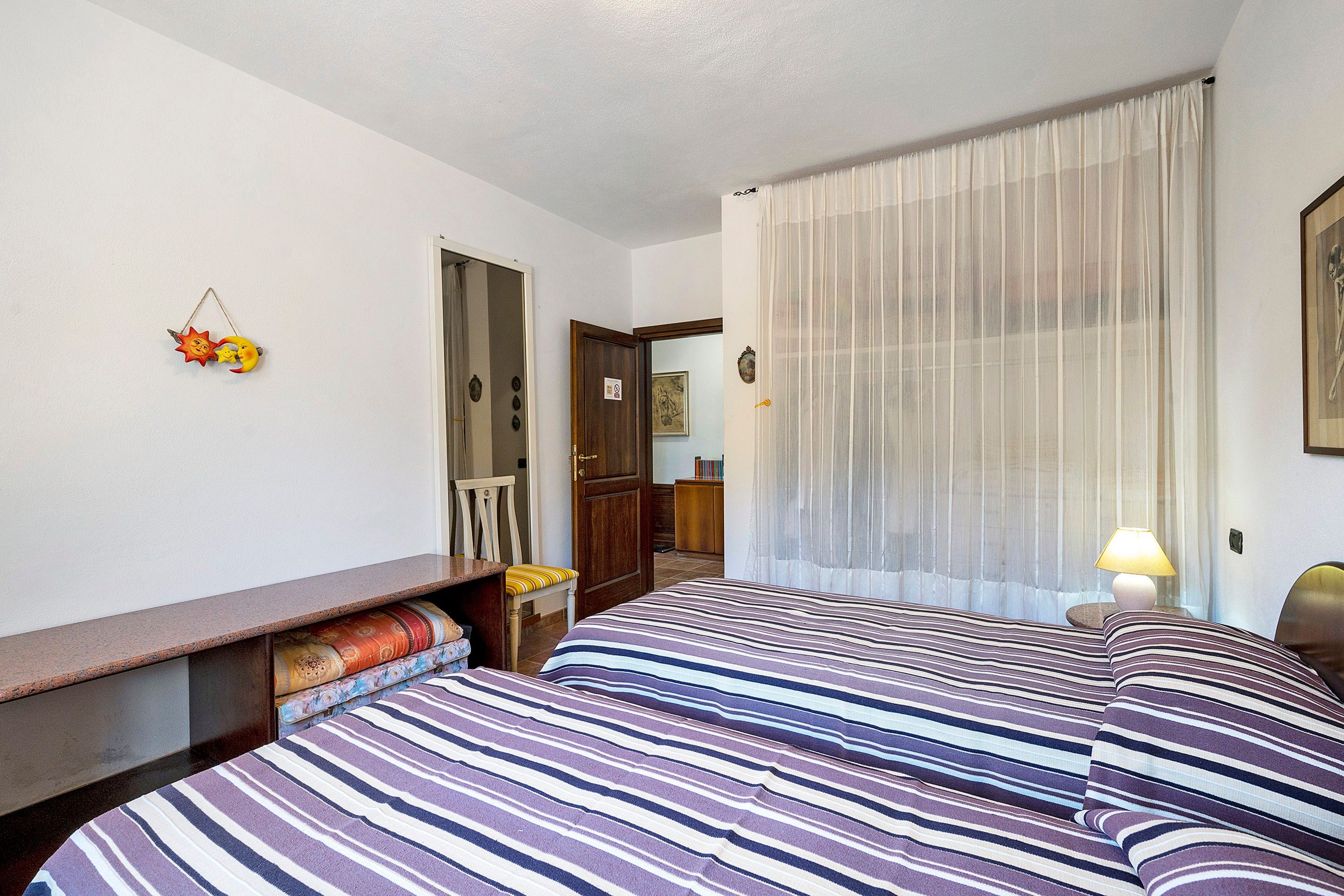A hotel room with two beds, a desk, a chair, a lamp, and a window with sheer curtains.