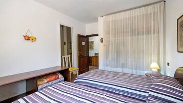 A hotel room with two beds, a desk, a chair, a lamp, and a window with sheer curtains.