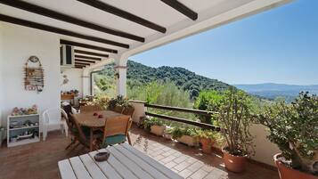 A covered outdoor patio with a table and chairs, surrounded by potted plants and a view of a hilly landscape.