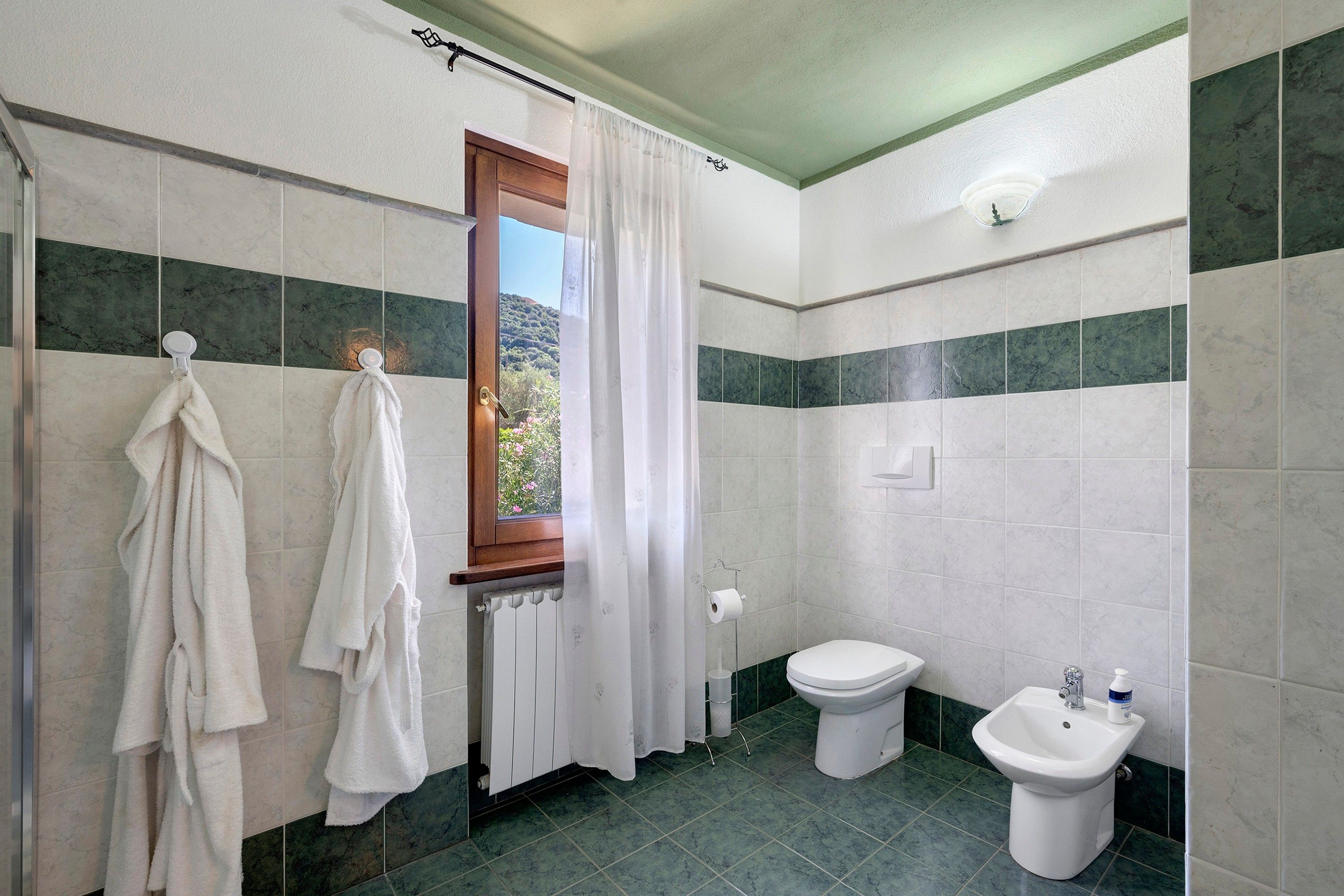 Ein Badezimmer mit Dusche, Toilette, Bidet und einem Fenster mit Vorhängen.