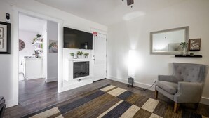 Un salon avec une cheminée, un fauteuil gris, un tapis à motifs et un miroir au mur.
