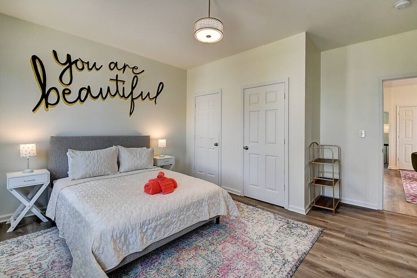 Une chambre à coucher comprenant un lit, deux tables de chevet, une lampe fixée au mur, une porte, un miroir et un couloir menant à une autre pièce.