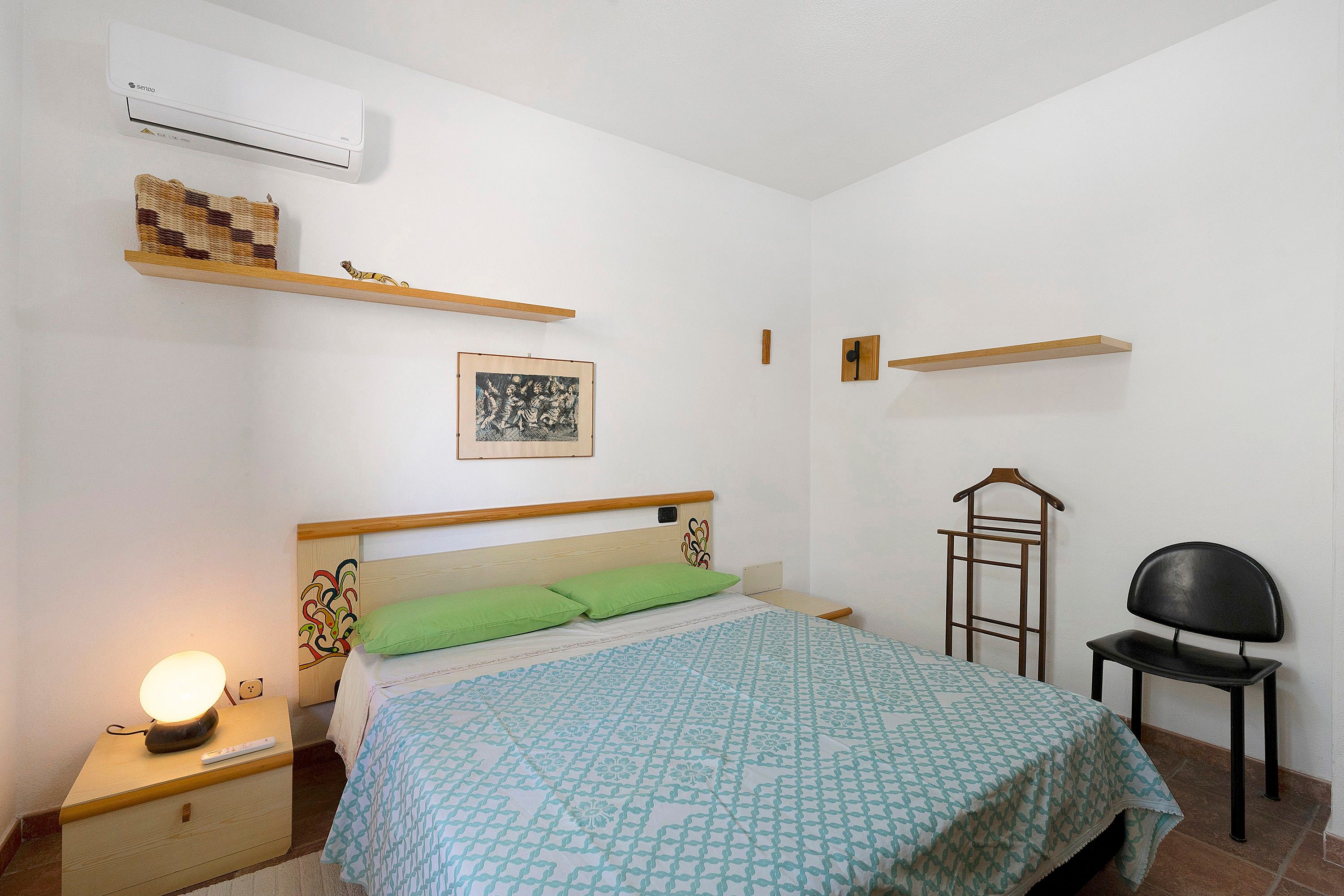Una camera da letto con un letto, un comodino, una lampada, mensole a parete, una sedia e un'opera d'arte appesa.