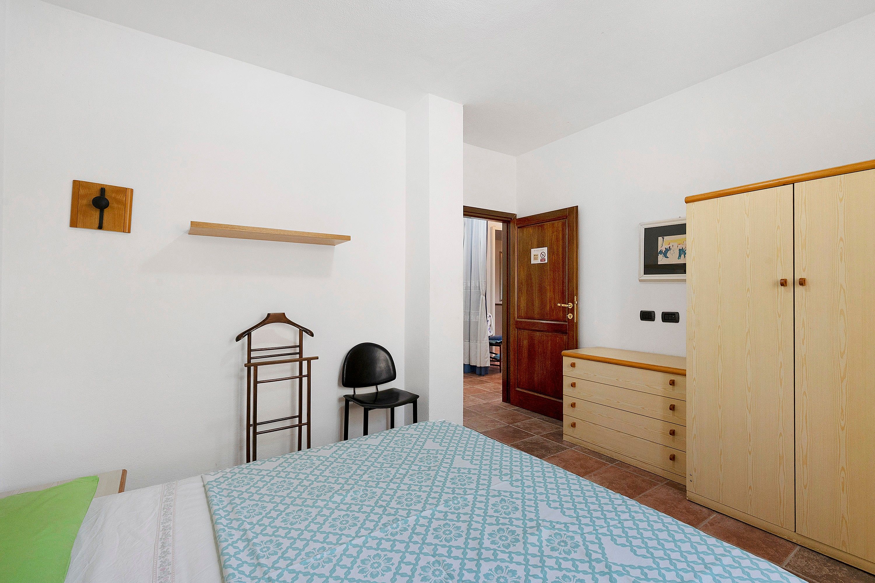 Ein Schlafzimmer mit einem Bett, einem Sessel, einem Kleiderschrank, einem Regal und einer Tür zu einem anderen Raum.