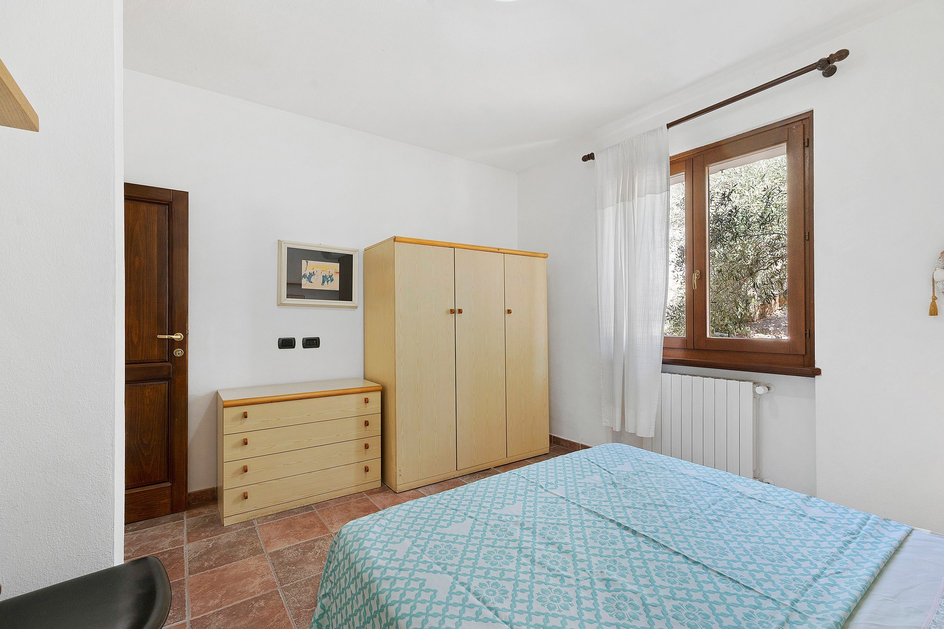 Ein Schlafzimmer mit Bett, Kleiderschrank, Kommode, einem Fenster mit Vorhängen und einer Heizung.