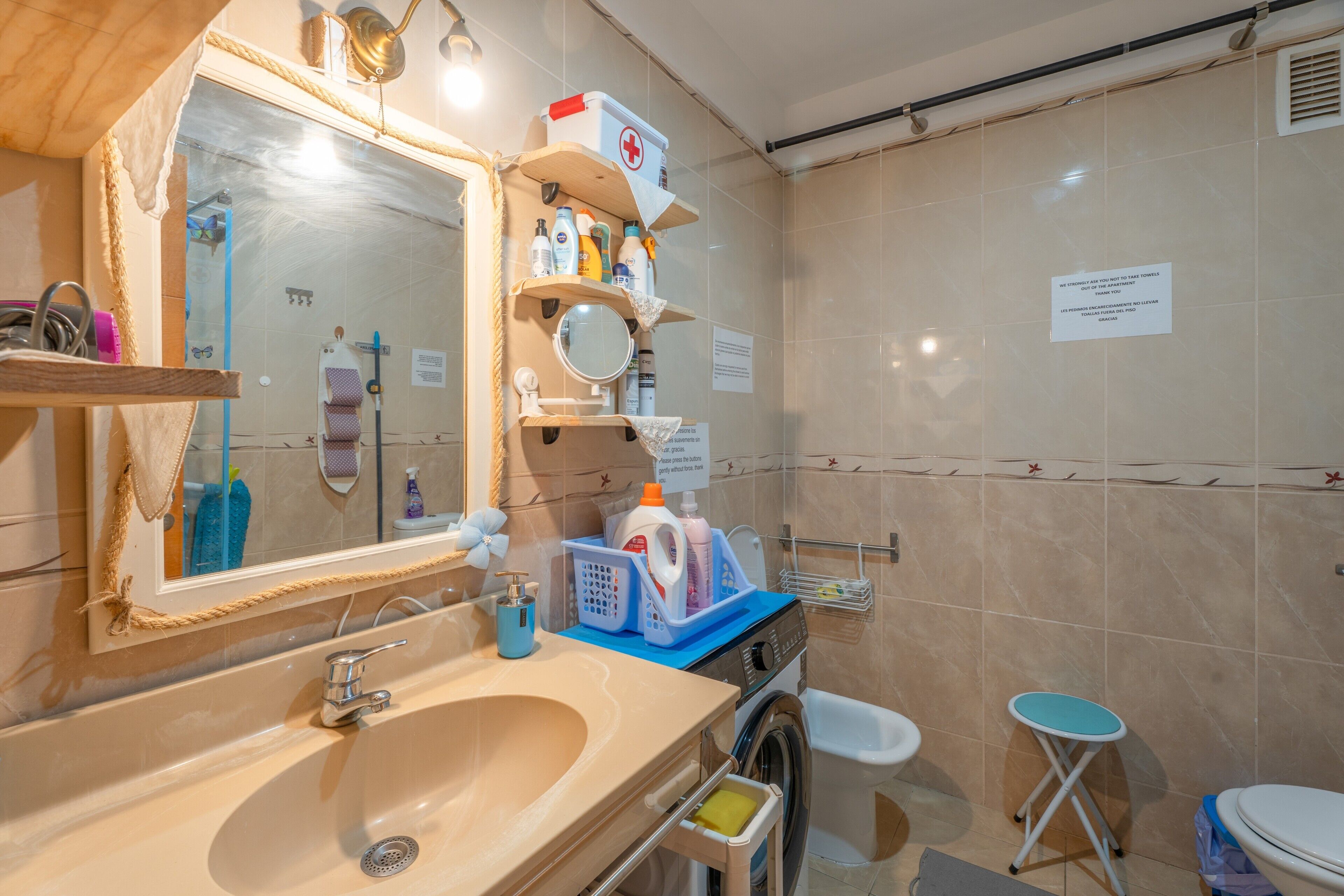 Un baño con lavamanos, espejo, estantes con artículos de aseo, lavadora e inodoro.