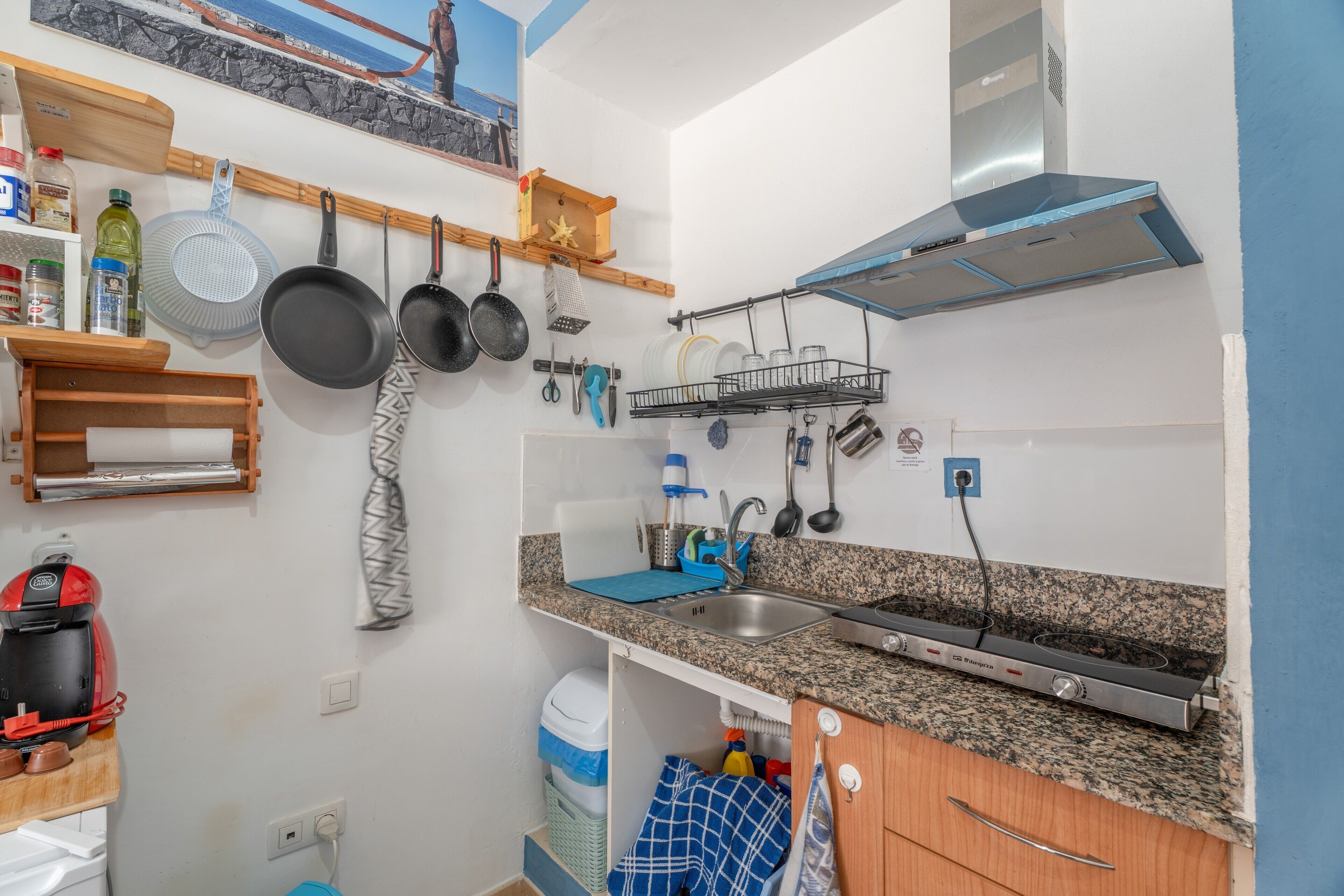 A kitchen with granite countertops, a stainless steel sink, hanging pots, a coffee maker, and a built-in range hood.