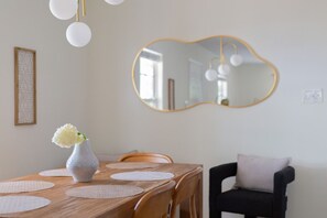 A dining area with a wooden table, white placemats, a vase with flowers, a mirror with a gold frame, and a black chair with a cushion.