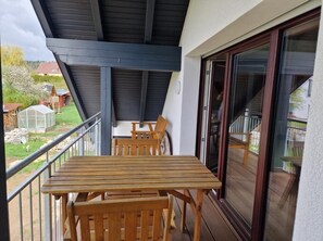 A balcony with a wooden table and chairs, a glass door, and a view of a garden and buildings.